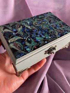 a hand holding an ornately decorated box with blue flowers on the outside and green leaves on the inside