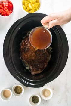 a person pouring sauce on top of a cooked meat in a slow cooker next to bowls of vegetables and seasonings