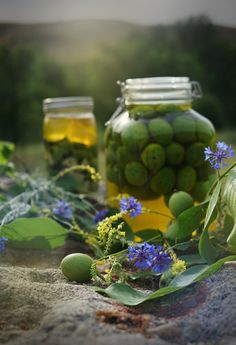 two jars filled with green olives and blue flowers on top of a stone surface