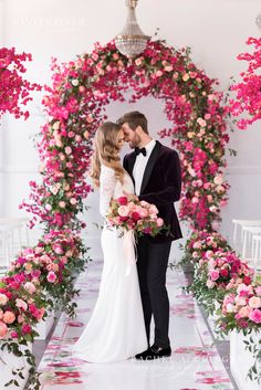 a bride and groom standing in front of an archway with pink flowers on the floor