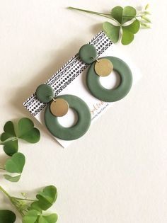 two green and gold earrings sitting on top of a white table next to some leaves