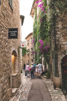 people are walking down an alley way with flowers growing on the walls and stone buildings