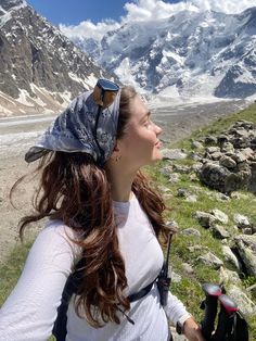 a woman standing on top of a mountain next to a lush green field with snow covered mountains in the background