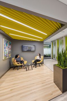 two people are sitting in yellow chairs near a plant on the wall and another person is standing