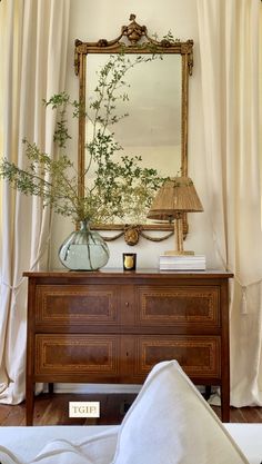 a dresser with a mirror, lamp and plants on it in front of a curtained window