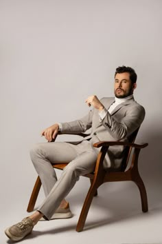 a man in a gray suit sitting on a wooden chair with his hand up to his chin