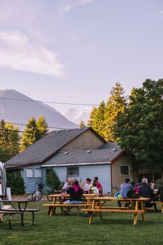 people sitting at picnic tables in front of a house with mountains in the background on a sunny day