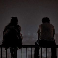 two people are sitting on a fence looking at the city lights in the foggy night