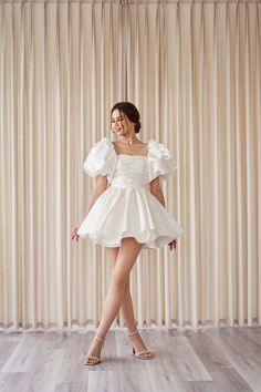 a woman standing in front of a curtain wearing a short white dress with ruffled sleeves