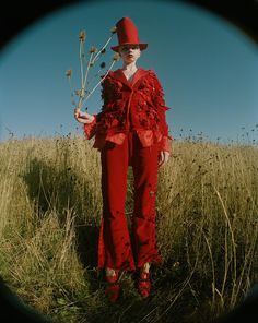 a woman standing in a field holding a flower and wearing a red hat with flowers on it