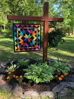 a colorful quilt is hanging on a cross in the garden with flowers and rocks around it