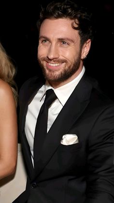 a man in a black suit and white shirt smiling at the camera with his hand on his lapel