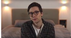 a man with glasses sitting in front of a bed looking at the camera while wearing a plaid shirt