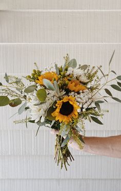 a person holding a bouquet of sunflowers and greenery in front of a white wall