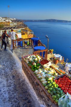 an outdoor fruit and vegetable market on the side of a cliff overlooking the ocean with people shopping