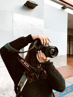 a woman taking a photo with her camera on the side of a building while wearing a black sweater