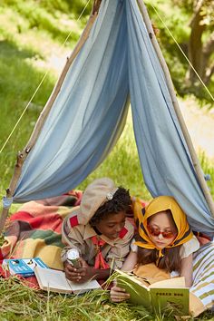 two children are laying on the grass under a teepee reading books and drinking tea