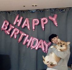 a woman holding two puppies in front of a happy birthday sign with pink balloons