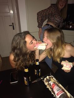 two young women are eating cake together at the kitchen table with beer bottles on the counter