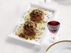 two meat patties with mushrooms and garnish on a white plate next to a glass of red wine