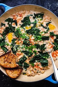 a pan filled with pasta and spinach on top of a blue table cloth next to two slices of bread