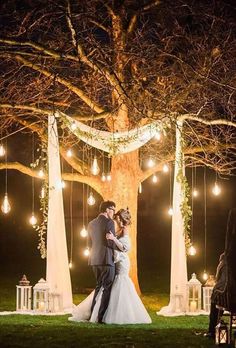 a bride and groom standing under a large tree with lights strung from it's branches