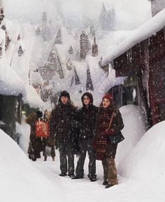 three children are standing in the snow near some houses and trees that are covered with snow