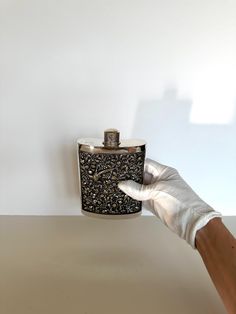 a hand in white gloves holding up a flask shaped like a black and gold flask