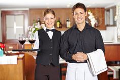 a man and woman holding a tray with two glasses of wine on it in front of a bar