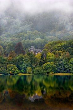 a house on the shore of a lake surrounded by trees and mist covered mountains in the background