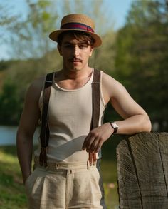 a young man wearing suspenders and a hat leaning on a fence post with his hands in his pockets