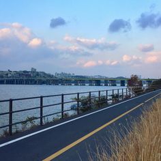 an empty road next to the water with a bridge in the background and grass on both sides