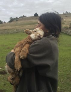 a woman holding a baby goat in her arms while standing on top of a lush green field