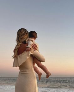 a woman in a dress holding a baby on the beach at sunset with waves crashing behind her