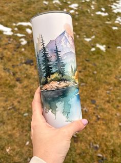 a person holding up a travel mug with mountains and trees painted on the side of it