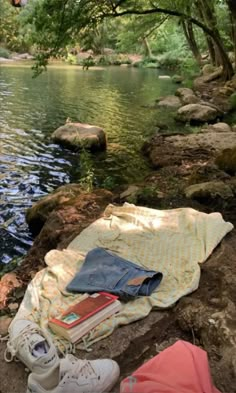 an open book and some books on a blanket by the water with trees in the background