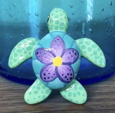 Clay Green sea turtle with purple flower on the shell Turtle Pottery Painting Ideas, Wooden Turtle Painted, Sea Turtle Clay Sculpture, Cute Animal Clay Sculptures, Cute Clay Turtle, Sea Turtle Ceramics, Turtle Ceramics Ideas, Painted Turtle Shell, Turtle Clay Art