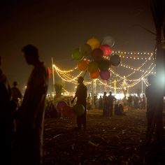 many people are walking around with balloons in the air and lights on the ground behind them