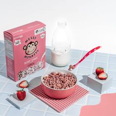 a pink bowl filled with cereal next to a box of milk and strawberries on a table