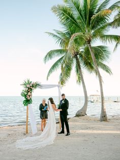 a bride and groom standing under a palm tree on the beach during their wedding ceremony