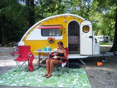 a woman sitting in a chair next to a yellow and white camper on the ground