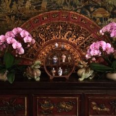 some pink flowers are sitting on top of a wooden dresser with an ornate fan in the background