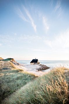 a house on the beach with grass and water in the foreground, under a partly cloudy sky