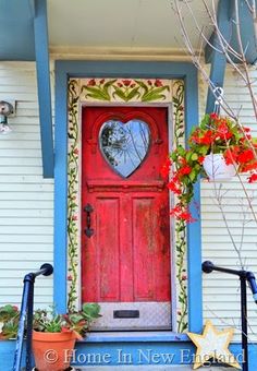 a red front door with a heart shaped mirror on it's side and potted plants next to it