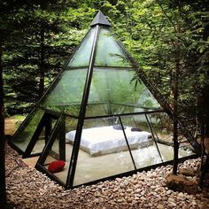 a bed sitting in the middle of a forest under a glass pyramid shaped tent that is surrounded by rocks and trees