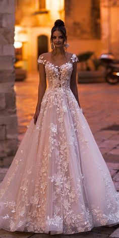 a woman wearing a wedding dress with flowers on the skirt and off the shoulder, standing in