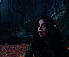 a woman with dark hair and green makeup looks at the camera while sitting in leaves on the ground