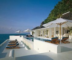 an outdoor swimming area with lounge chairs and umbrellas on the deck overlooking the ocean