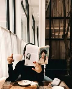 a woman sitting at a table reading a book and holding a coffee cup in front of her face