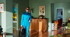 a woman in a blue suit and sunglasses is holding a black handbag while standing in a living room